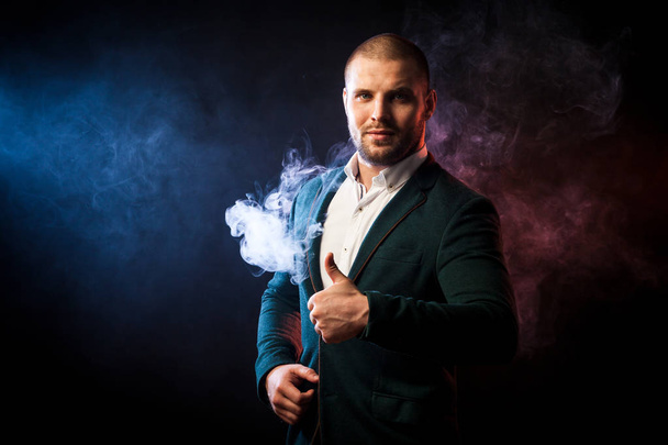 Young handsome businessman man in white shirt and green jacket smiling and showing thumb up opposite red and blue smoke from wipe on black isolated background - 写真・画像