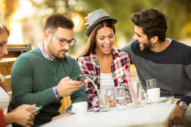 Grupo de cuatro amigos usando teléfonos inteligentes, divirtiéndose y tomando café juntos
.  - Foto, Imagen