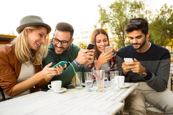 Grupo de cuatro amigos usando teléfonos inteligentes, divirtiéndose y tomando café juntos
.  - Foto, imagen