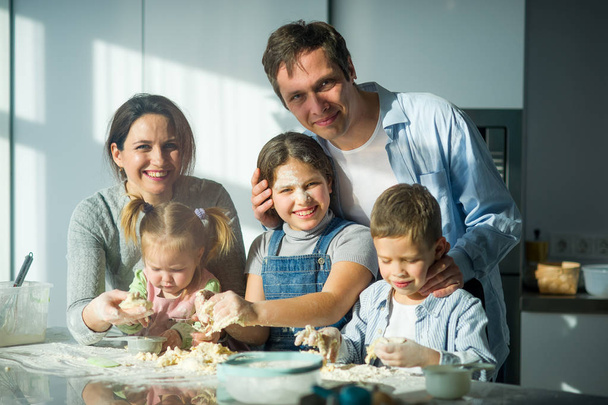 Большая семья готовит что-то из теста. Родители и трое детей за кухонным столом. Веселое и полезное времяпрепровождение
. - Фото, изображение
