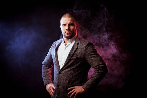 Young handsome man businessman in white shirt and  gray suit  stands and looks at the camera against blue and red smoke from a vape  on black isolated - Fotoğraf, Görsel