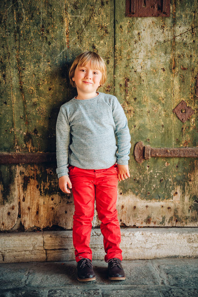 Szép kis fiú visel piros chinos és világos kék pulóver, elegáns gyerek jelentő vintage régi falhoz szabadtéri portréja. Divat a kisgyermekek - Fotó, kép