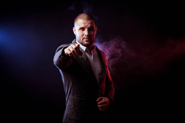 Young handsome man businessman in white shirt and  gray suit  show your index finger and choose you against red smoke from a vape  on black isolated  - Фото, изображение
