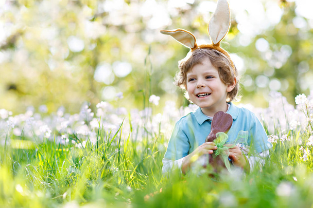 Χαριτωμένο αγοράκι με αυτιά πασχαλινού κουνελιού που γιορτάζει παραδοσιακή γιορτή. Ευτυχισμένο παιδί τρώει σοκολάτα λαγουδάκι fugure - Φωτογραφία, εικόνα