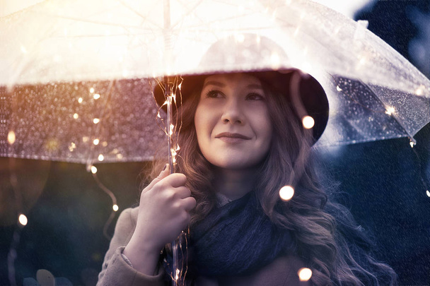 Young woman dreamy face standing under rain with illuminated transparent umbrella and smiling - Фото, зображення