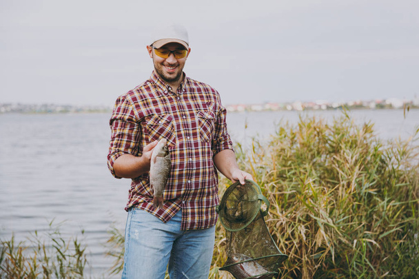 Young unshaven smiling man in checkered shirt, cap, sunglasses keeps in hands green fishing grid and fish which he caught on shore of lake near shrubs and reeds. Lifestyle, fisherman leisure concept - Photo, Image