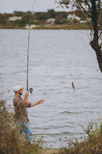 Молодой небритый мужчина в клетчатой рубашке, кепке и солнечных очках вытаскивает удочку с пойманной рыбой на озере с берега возле кустарников и тростника. образ жизни, отдых, рыбацкая концепция досуга
 - Фото, изображение