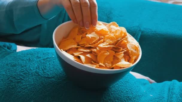 Married couple lies on a sofa under a blue blanket and eats potato chips from a plate that stands on the big belly of a man. Unhealthy food - Footage, Video