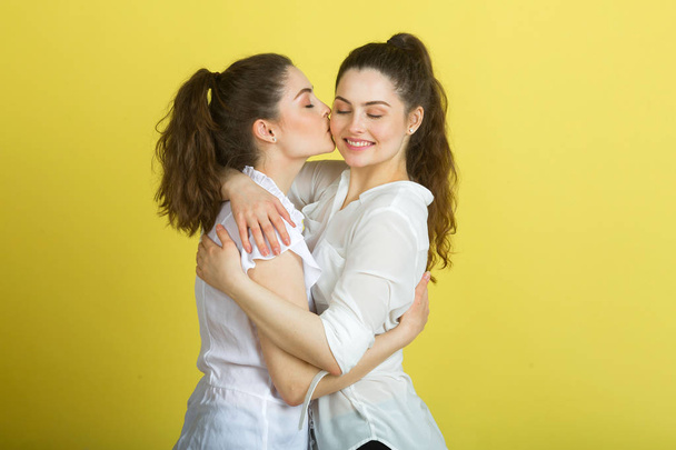 two beautiful joyful sisters twin girls in white blouses on a yellow background hugging each other and kissing - Foto, immagini