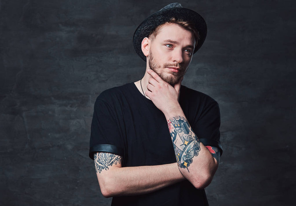 Portrait of handsome bearded hipster with crossed tattooed arms, dressed in a black t-shirt and hat. Isolated on a dark background. - Photo, Image