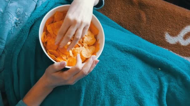 Vrouw is liggend op een bank onder een blauwe deken en chips eten - Video