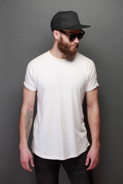 Hipster handsome male model with beard wearing white blank t-shirt with space for your logo or design over gray background - Фото, изображение