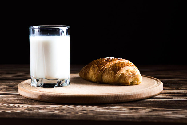 Um delicioso croissant em uma placa redonda de madeira perto de um copo de leite em um fundo preto
. - Foto, Imagem