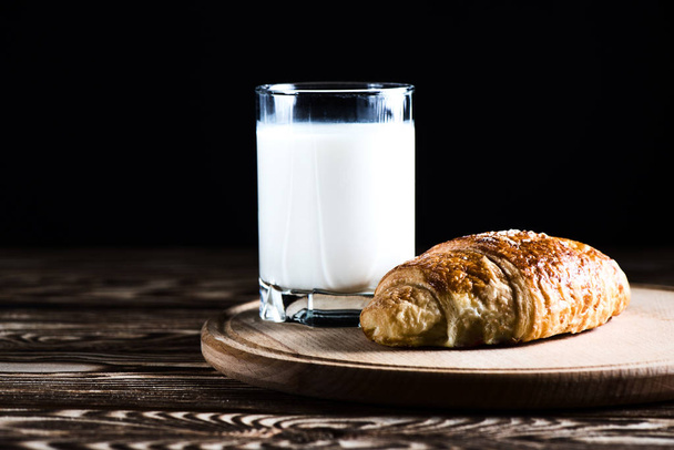 A delicious croissant on a round wooden plate near a glass of milk on a black background. - Photo, Image