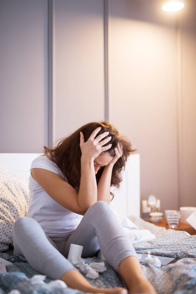 несчастная больная женщина сидит на кровати и с помощью планшета ПК, пытаясь работать дома страдает от гриппа
  - Фото, изображение