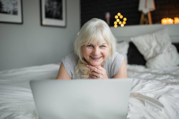Happy senior lady lying on bed using notebook - Фото, изображение