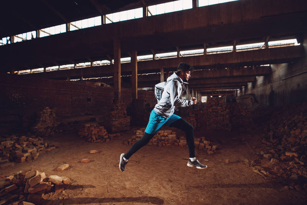 portrait of motivated afro-american man running in abandoned hangar place with earphones - Photo, Image
