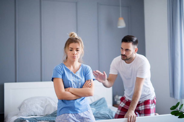 young angry blonde woman sitting on the bed, her serious boyfriend behind. Young unhappy couple arguing in bedroom - Zdjęcie, obraz