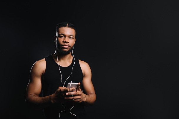 young african american sportsman in earphones holding smartphone and looking at camera isolated on black - Fotoğraf, Görsel