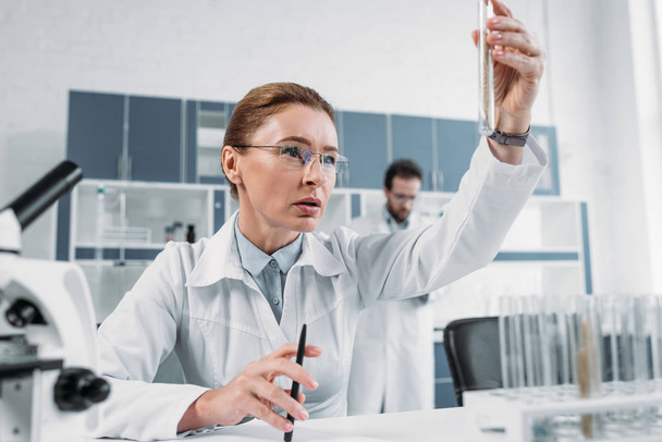 selectieve aandacht van vrouwelijke wetenschapper op zoek naar buis met reagens in de hand met collega achter in lab - Foto, afbeelding