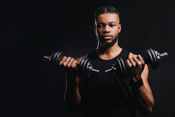 handsome young sportsman holding dumbbells and looking at camera isolated on black  - Photo, Image