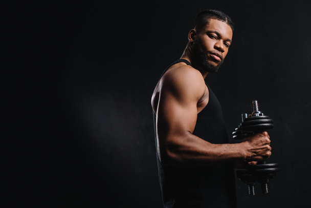 Мускулистый африканский спортсмен держит гантели и смотрит на камеру изолированную на черном
 - Фото, изображение