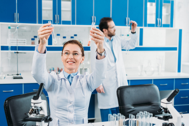 Selektywny fokus kobieta naukowiec patrząc na rurki z odczynnikami w ręce z kolega za w laboratorium - Zdjęcie, obraz