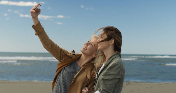two smiling young women making selfie on mobile phone on autumn day on beach - Foto, Bild