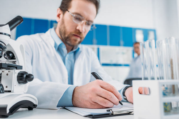 selective focus of scientist in white coat and eyeglasses making notes in notepad at workplace in laboratory - Foto, afbeelding