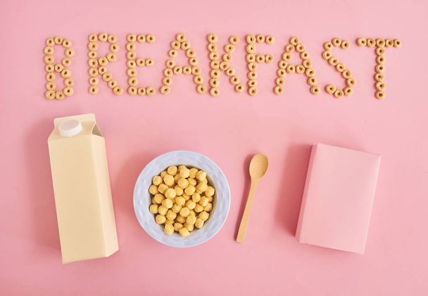 Placa de cereales o copos de maíz, botella de leche, cuchara, caja y palabra Desayuno sobre fondo rosa, vista superior
 - Foto, Imagen