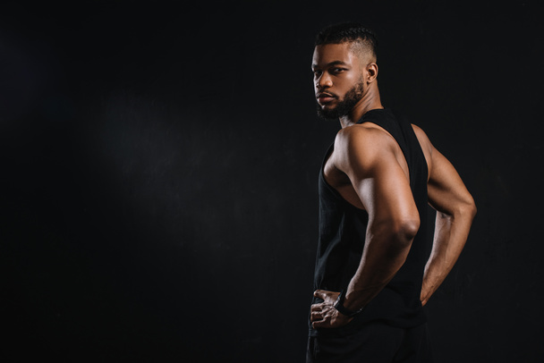 handsome young african american sportsman standing with hands on waist and looking at camera isolated on black - Fotografie, Obrázek