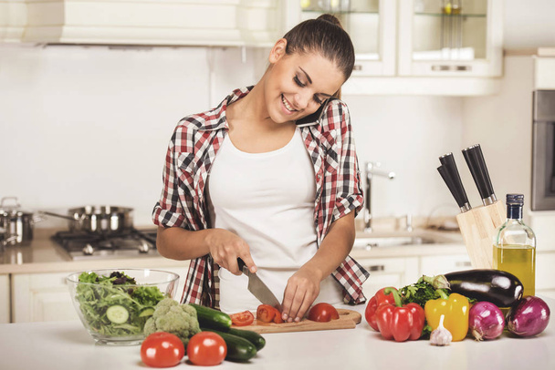 Young woman is chopping vegetables in the kitchen and speaking by phone - Foto, afbeelding
