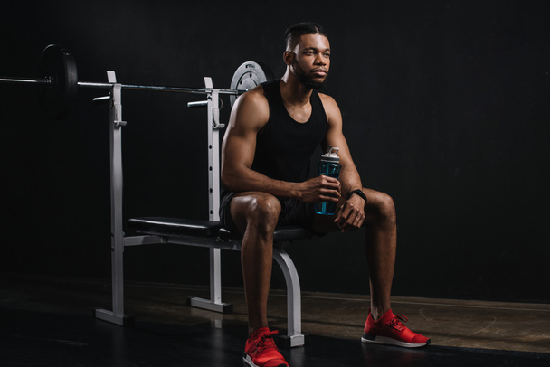 young african american sportsman holding bottle of water and looking away on black - Фото, изображение