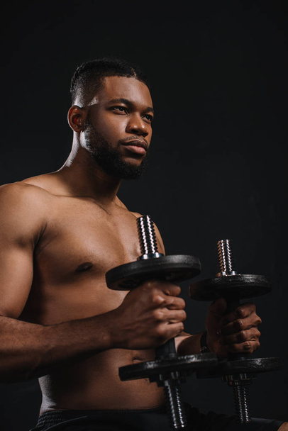 muscular shirtless young african american man holding dumbbells and looking away on black - Фото, зображення