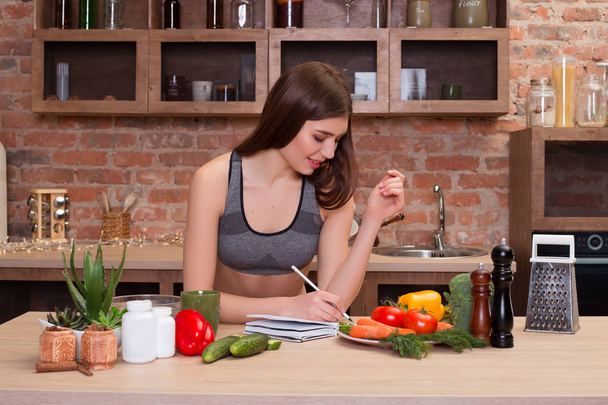 Zdrowie to skarb! Młoda atrakcyjna kobieta uśmiechający się otoczony jest zdrowy warzyw siedzi przy stole w kuchni i pisze w swoim pamiętniku wszystko o zdrowym odżywianiu. - Zdjęcie, obraz