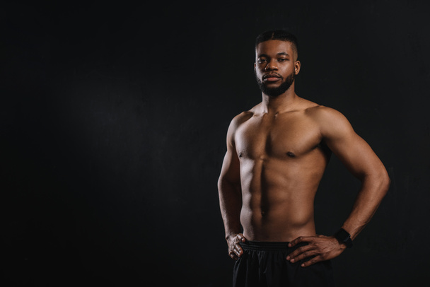 muskulöser, hemdloser afrikanisch-amerikanischer Mann, der mit den Händen auf der Taille steht und isoliert auf schwarz in die Kamera schaut - Foto, Bild