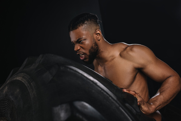 vista lateral de la musculosa joven afroamericano deportista levantar neumático en negro
 - Foto, Imagen