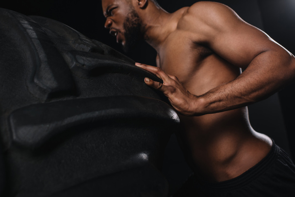 low angle view of muscular young african american sportsman lifting tire on black - Foto, Bild