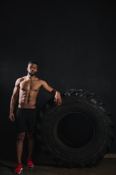 mięśni shirtless młodego sportowca african american opierając się o opony i patrząc na kamery na czarny - Zdjęcie, obraz