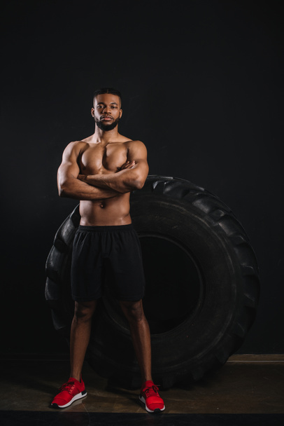 visão de comprimento total do atleta afro-americano sem camisa de pé com braços cruzados perto de pneu grande e olhando para a câmera em preto
  - Foto, Imagem