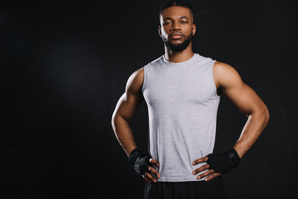 handsome young african american sportsman standing with hands on waist and looking at camera isolated on black - Foto, imagen