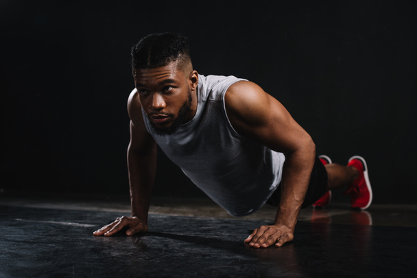 young muscular african american man in sportswear doing push-ups and looking away on black - Фото, зображення