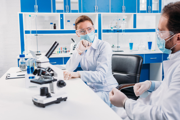 επιστημονικούς ερευνητές σε ιατρικές μάσκες και γυαλιά έχοντας συζήτηση στο χώρο εργασίας με μικροσκόπιο στο εργαστήριο - Φωτογραφία, εικόνα