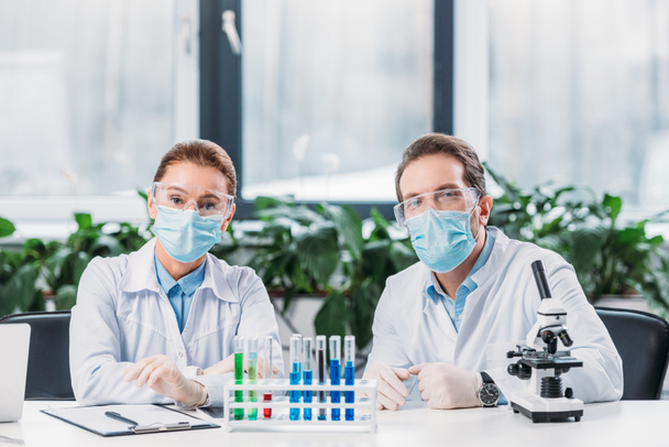 pesquisadores científicos em óculos e máscaras médicas sentados no local de trabalho com reagentes em tubos e microscópio em laboratório
 - Foto, Imagem