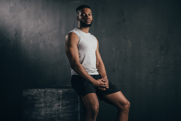 young athletic african american man in sportswear sitting and looking at camera on black  - Foto, imagen