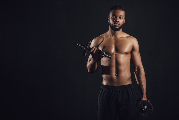 muskulöser, hemdloser afrikanisch-amerikanischer Sportler, der mit Kurzhanteln trainiert und isoliert auf schwarz in die Kamera schaut   - Foto, Bild