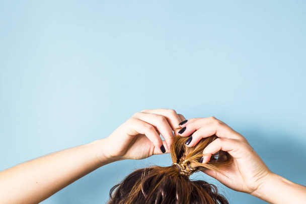 The woman straightens the disheveled bun on her head with her hands with a black manicure. Dark hair is tied with a transparent spiral elastic band. Modern fast hairstyle. Blue background. Copyspace - Photo, Image