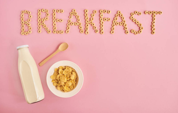 Deska z obilovin nebo kukuřičné lupínky, láhev mléka a slovo snídaně na růžovém pozadí, pohled shora. Mockup design s prostorem pro vstup textu - Fotografie, Obrázek