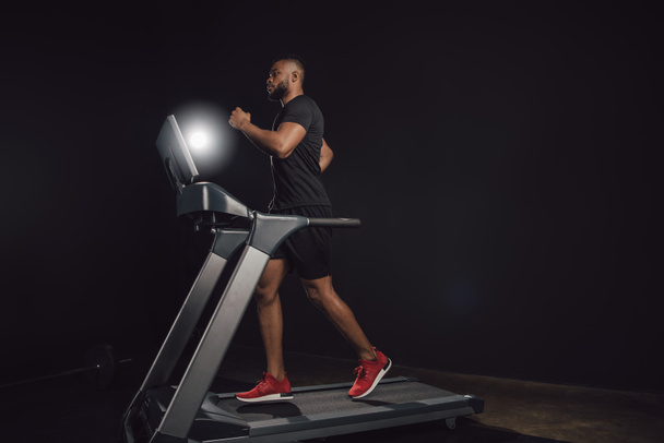 side view of athletic young african american man running on treadmill on black - 写真・画像