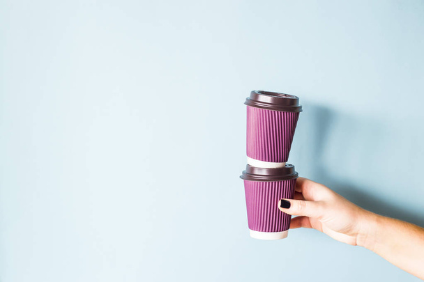 Purple paper kraft disposable cups for coffee with plastic lids in a female hand. One glass stands on the other. Tower of coffee. On blue background - Photo, Image